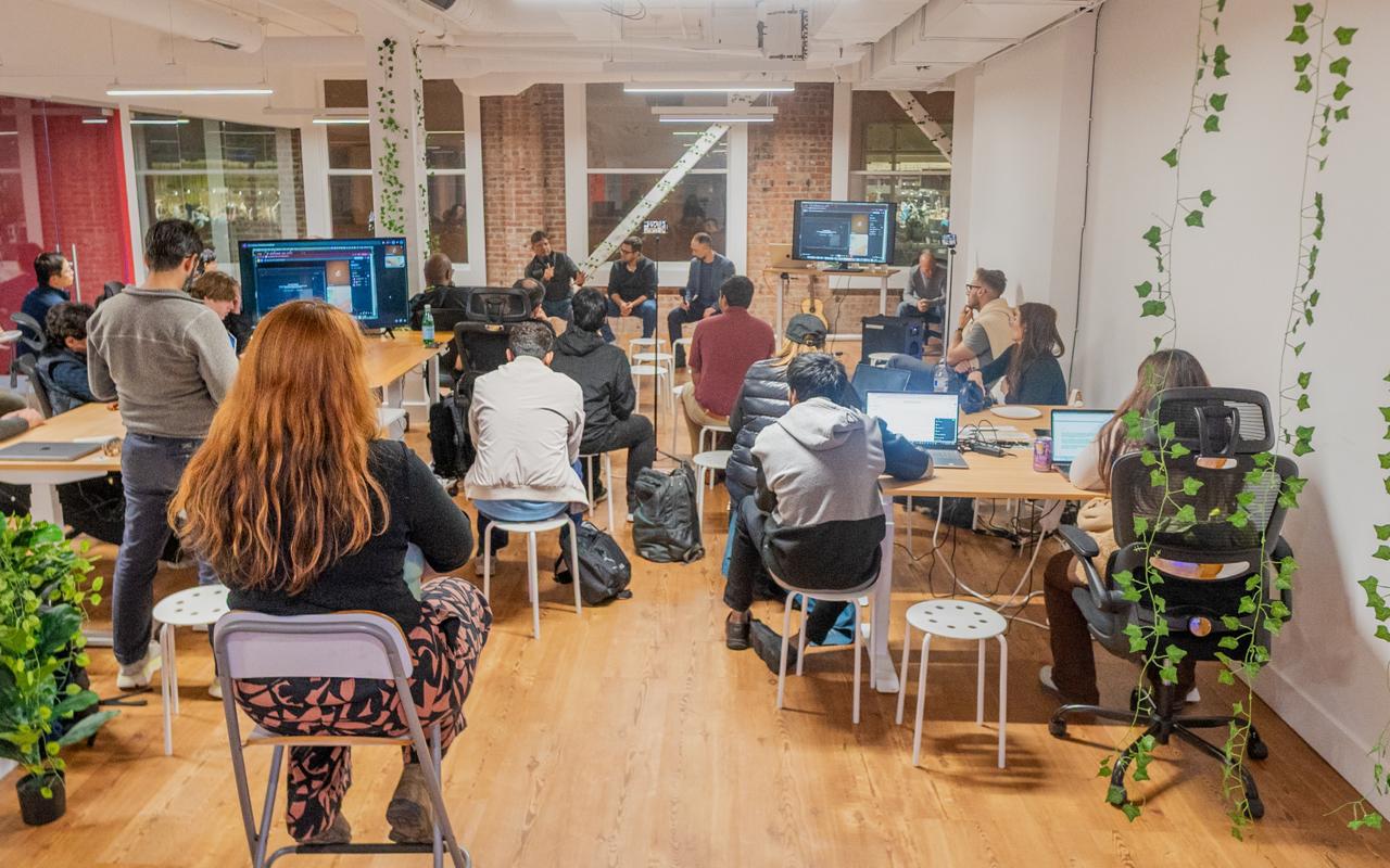 Full club meeting overhead shot — laptops, multiple screens with code, brick interior and hanging plants